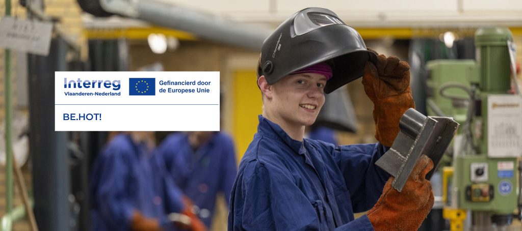Student in overall en laskap glimlacht in een werkplaats tijdens een praktijkles over duurzaam bouwen, onderdeel van het Interreg-project Building Eco Heroes of Tomorrow (BE.HOT)