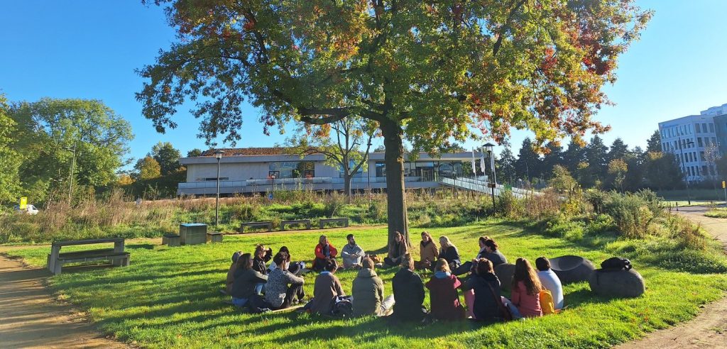 Een groep mensen zit in een kring onder een grote boom in het zonlicht, tijdens een bijeenkomst over natuurinclusief onderwijs binnen de Community of Practice.
