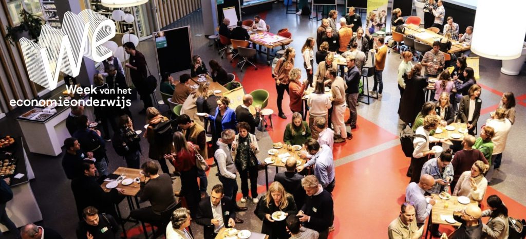 Drukke netwerkbijeenkomst tijdens de Week van het Economieonderwijs, met deelnemers die in gesprek zijn en samen eten in een grote ruimte.