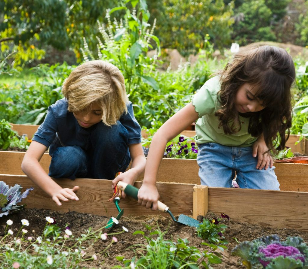 Twee jonge kinderen werken samen in een moestuin en planten bloemen, symbool voor leren door te doen en zorg voor de natuur.