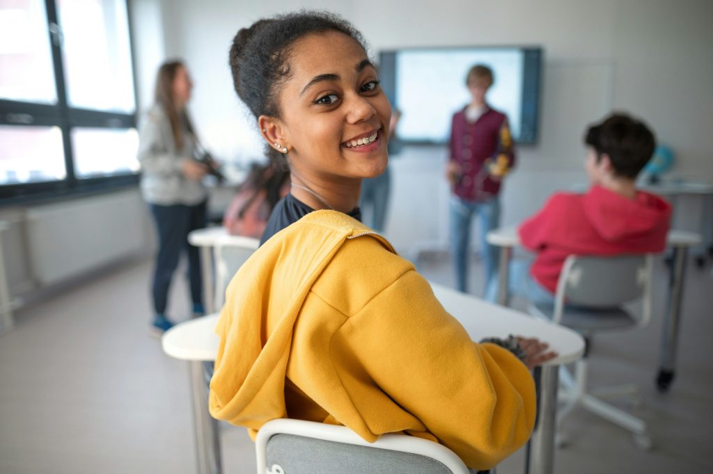 Leerling kijkt lachend om in de klas terwijl klasgenoten samenwerken aan een opdracht, symbool voor betrokken en toekomstgericht onderwijs.