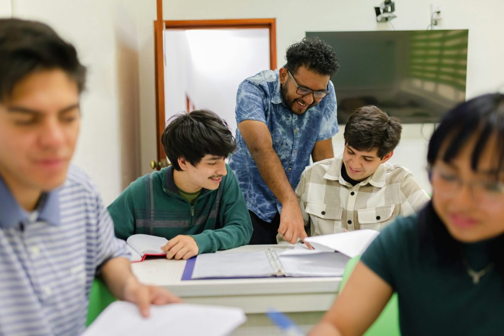 Docent begeleidt twee leerlingen tijdens de les en wijst iets aan in hun werkmap, symbool voor inspirerend en betrokken onderwijs.